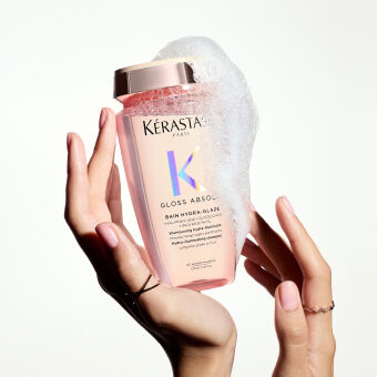 Flacone di shampoo Kerastase Gloss Absolu tra le mani, con schiuma bianca che si forma sopra il tappo, su sfondo chiaro.