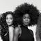 A black-and-white portrait of three women standing side by side, each showcasing different hairstyles: one with loose curls, one with a large afro, and one with wavy red hair. They are wearing sleeveless black tops and are posed against a plain background.