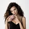 A woman with curly hair poses in a black outfit while holding a pink bottle of hair product close to her face, looking confidently at the camera.