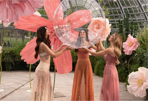 Tre donne in abiti fluidi interagiscono con bolle trasparenti e fiori giganti rosa in un ambiente lussureggiante.