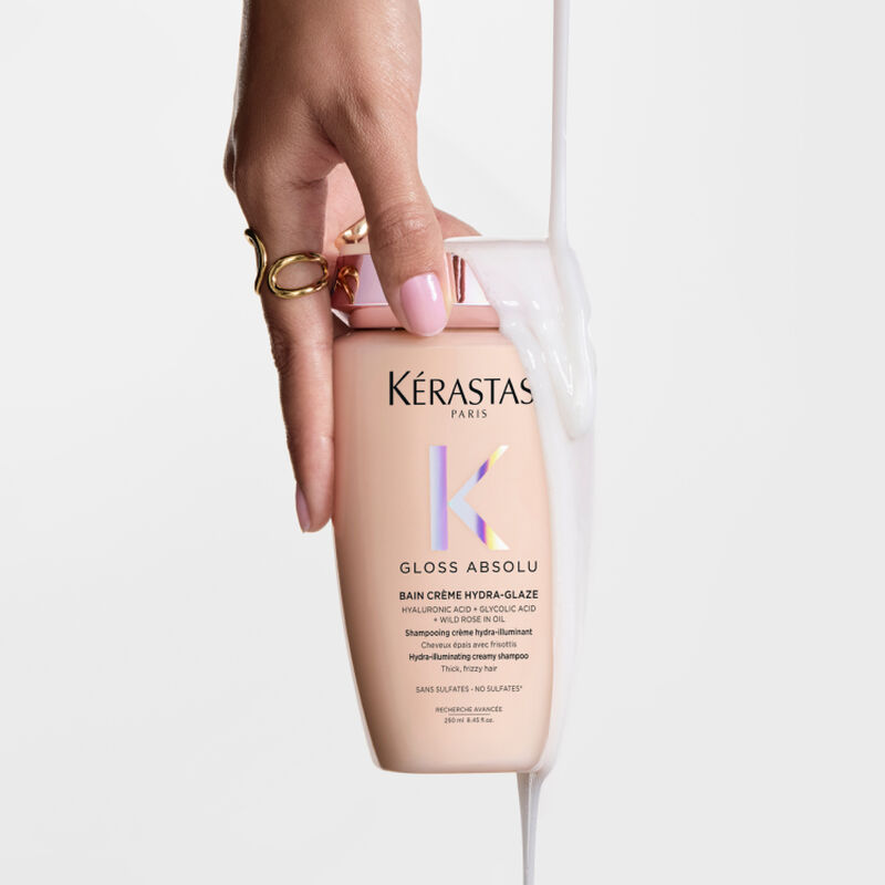 Mano che tiene un flacone di shampoo Kerastase Gloss Absolu con un liquido bianco cremoso che scorre sopra.