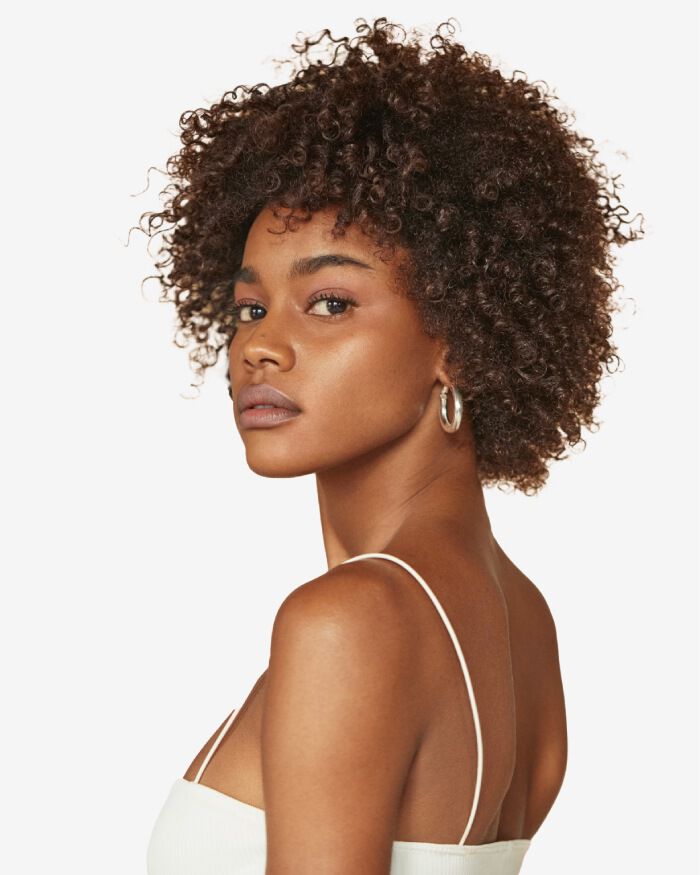 Ritratto di una giovane donna con capelli ricci e voluminosi, che guarda oltre la spalla, indossando una canotta bianca.