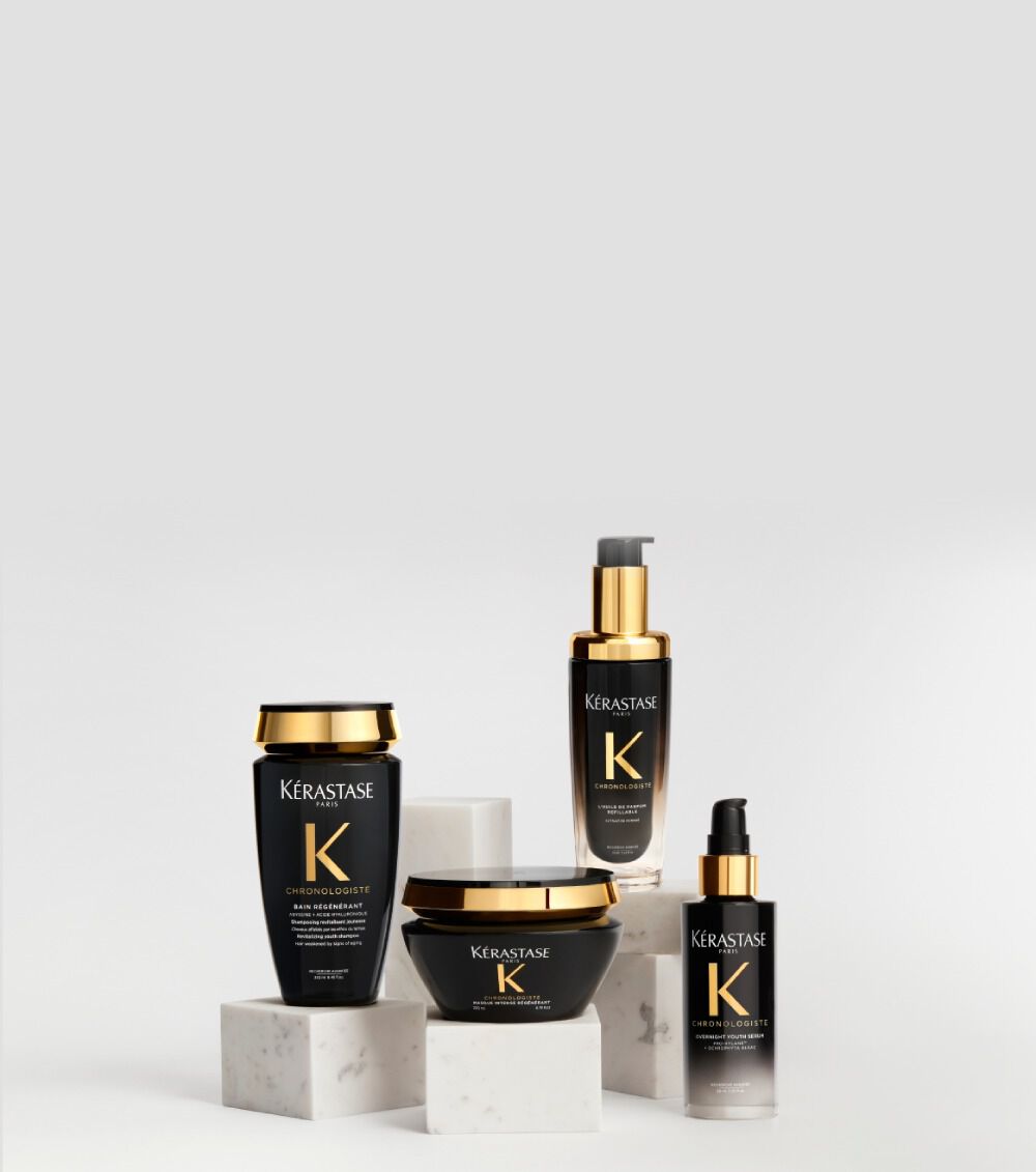 Prodotti per capelli della linea Kerastase Chronologiste in flaconi neri e oro, elegantemente disposti su basi di marmo bianco su uno sfondo chiaro.