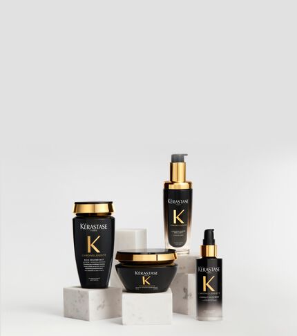 Prodotti per capelli della linea Kerastase Chronologiste in flaconi neri e oro, elegantemente disposti su basi di marmo bianco su uno sfondo chiaro.