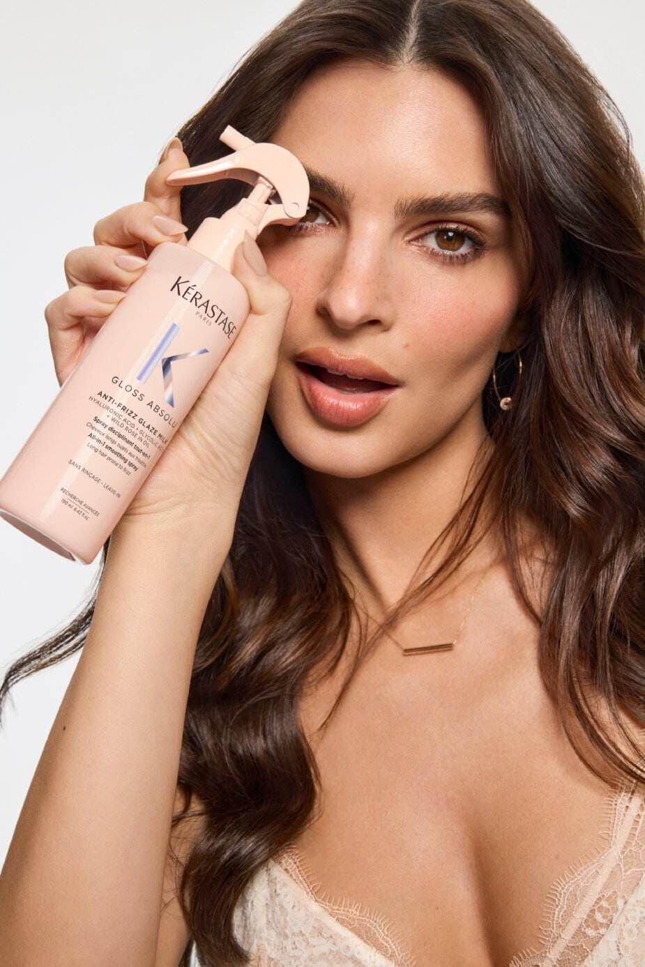 Una donna con lunghi capelli castani tiene in mano una bottiglia di prodotto per capelli Kerastase, sorridendo verso la fotocamera. Indossa un top elegante e orecchini dorati.