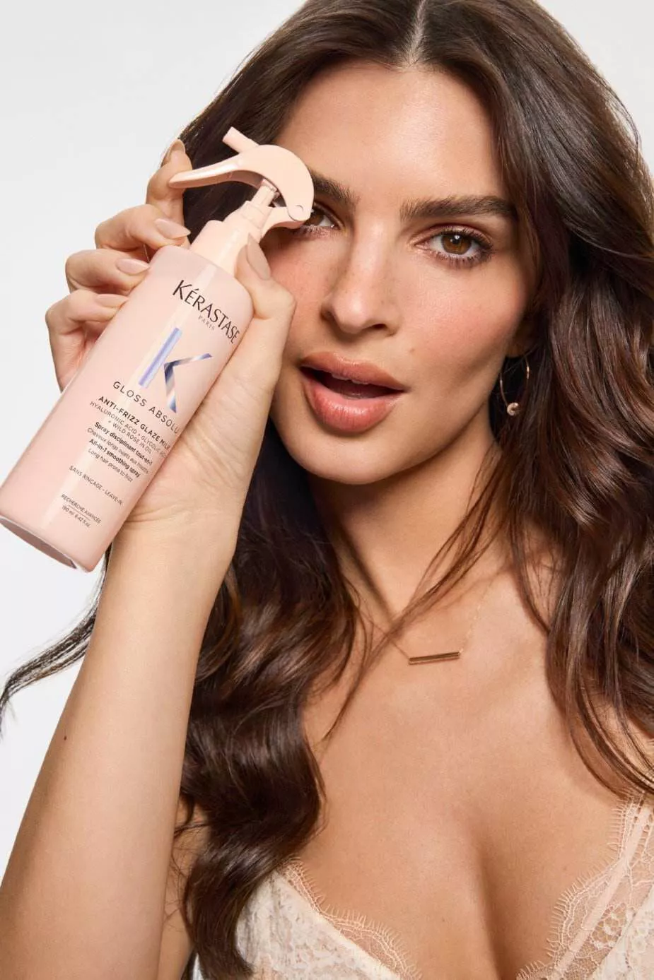 Una donna con lunghi capelli castani tiene in mano una bottiglia di prodotto per capelli Kerastase, sorridendo verso la fotocamera. Indossa un top elegante e orecchini dorati.