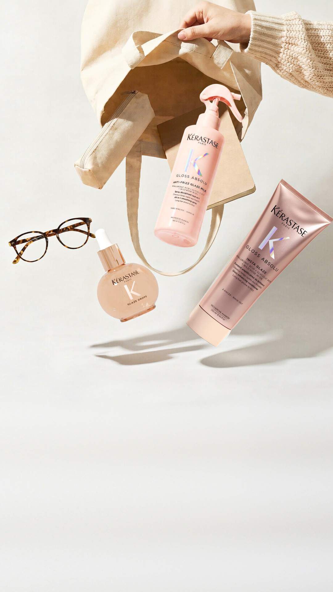 Tre flaconi di prodotti per capelli Kerastase (Elixir Ultime, Genesis e Nutritive) sono disposti su una superficie bianca accanto a una macchina fotografica vintage e a diverse peonie bianche con foglie verdi. Il flacone dorato Elixir Ultime e a sinistra con una pompa nera, il flacone rosa Genesis con un contagocce argentato e al centro, e il flacone sfumato rosso-arancio Nutritive con un contagocce argentato e a destra. La macchina fotografica argentata con il suo laccetto nero e a sinistra dei prodotti.