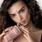 A woman with long, wavy hair is holding a pink skincare product close to her face, looking directly at the camera with a confident expression. She is wearing a simple black top against a light background.