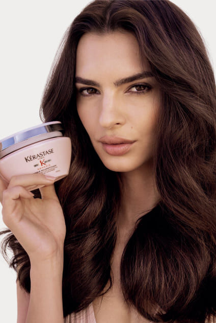 Donna che tiene un vasetto rosa della maschera Kerastase Genesis Masque Reconstituant, che presenta il prodotto.