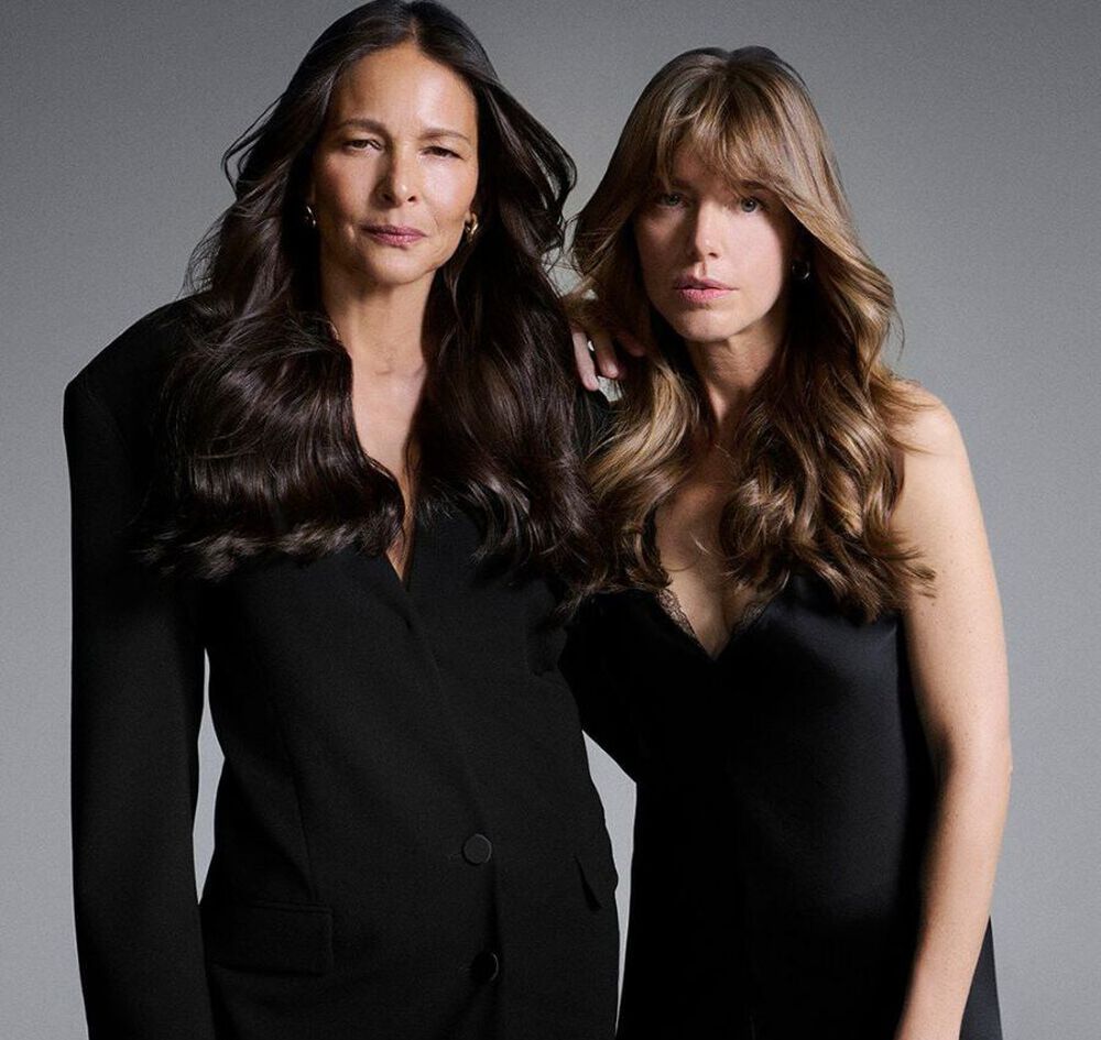 Due donne in posa insieme in eleganti abiti neri, che mostrano i loro capelli lunghi e sani.