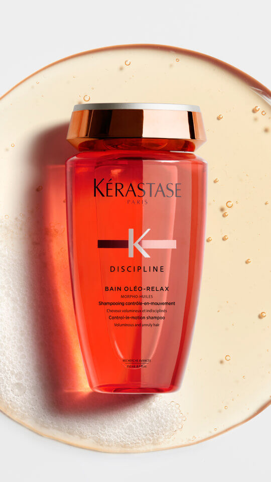 A bottle of Kerastase Discipline Bain Oleo-Relax shampoo, featuring a sleek design in a vibrant orange hue, set against a circular splash of water.