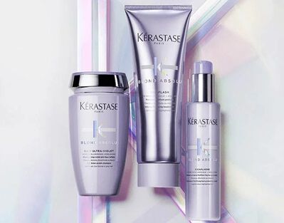 Three Kerastase hair care products are displayed against an iridescent background. The products include a jar, a tube, and a spray bottle, all from the "Blond Absolu" line, featuring a soft purple color scheme.