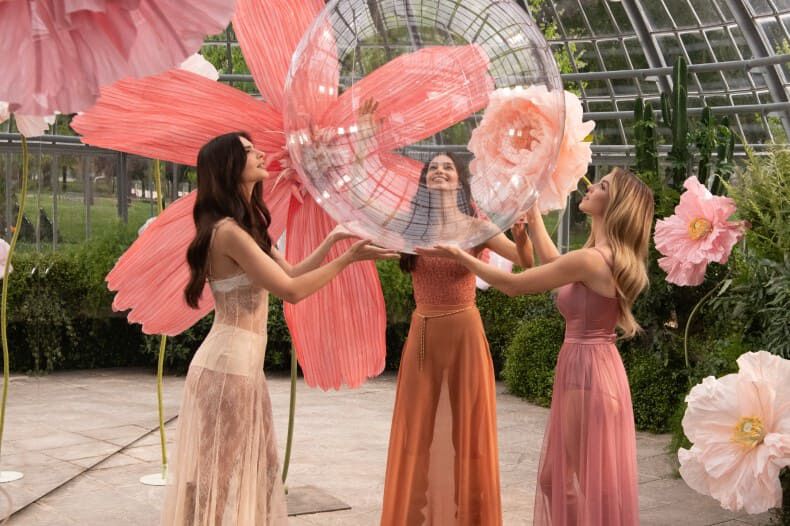 Three women in a botanical setting are joyfully interacting with a large, transparent orb surrounded by vibrant flower decorations.
