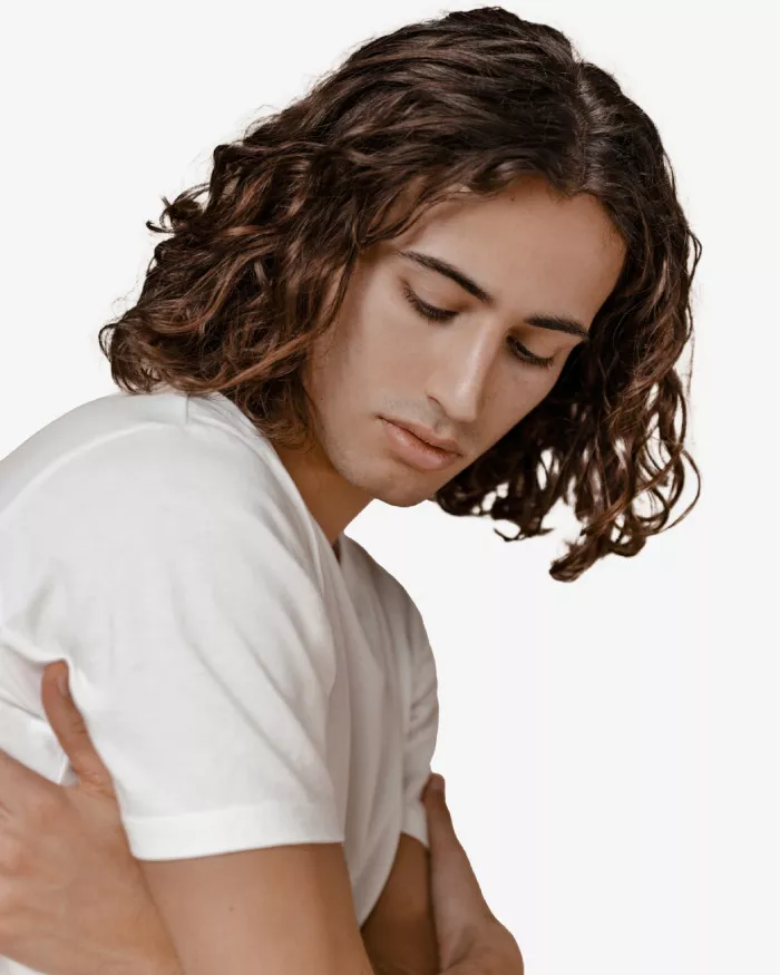 Un giovane uomo con i capelli ricci e lunghi, indossa una maglietta bianca e ha uno sguardo pensieroso mentre si autoabbraccia.