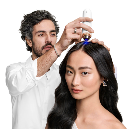 A stylist applies a hair treatment to a woman's scalp using a device, while she looks directly at the camera, showcasing her long, dark hair and minimalist makeup.
