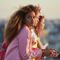 A woman with long, curly hair gazes thoughtfully into the distance, while two other individuals are seen in profile beside her against a sunset backdrop.
