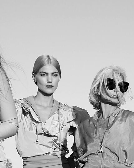 Two women stand outdoors, dressed in fashionable attire. One has sleek, blonde hair and wears a light-colored jacket, while the other sports short, wavy hair and oversized sunglasses. The image is in black and white, creating a dramatic contrast against the clear sky.