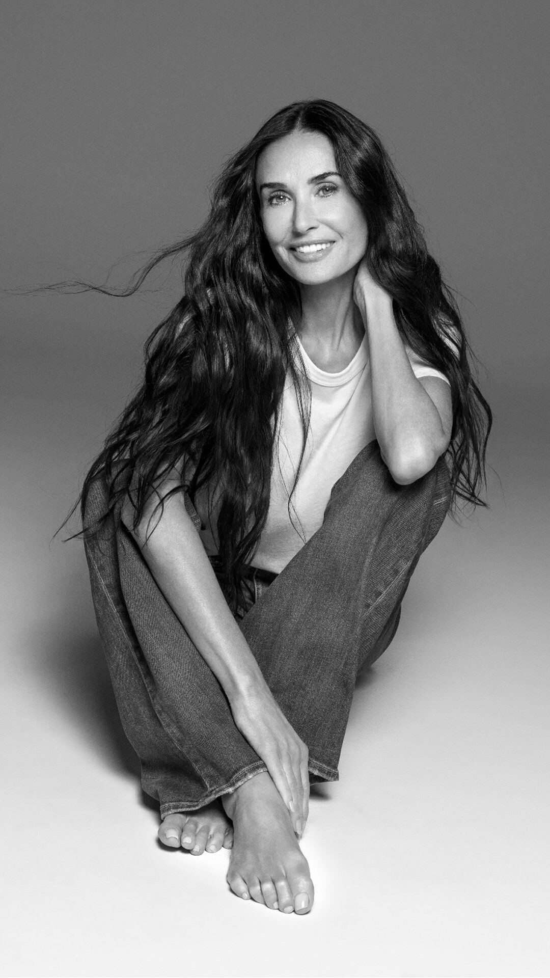 Demi Moore seduta a gambe incrociate con un grande sorriso radiante.