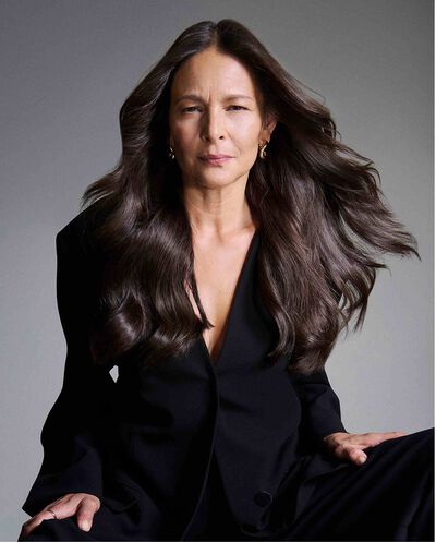 Ritratto di una donna matura con lunghi capelli castani voluminosi che indossa un blazer nero su sfondo grigio.