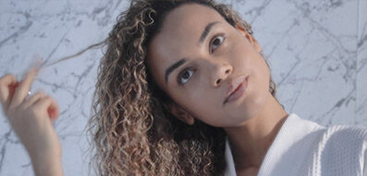 Una giovane donna con capelli ricci sorride mentre gioca con una ciocca di capelli, su uno sfondo di marmo chiaro.