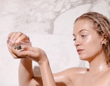 A person with wet hair holds a small jar in one hand, looking thoughtfully at the contents, in a marble bathroom setting.