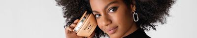 A young woman with curly hair holds a gold hair care product close to her face, smiling confidently against a white background.