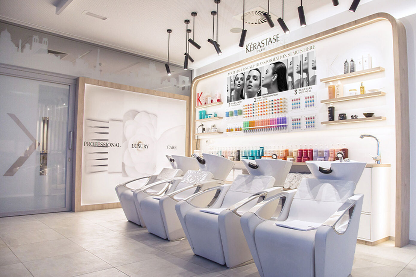 A modern hair salon interior featuring sleek white styling chairs in front of a wall display of colorful hair products. The space is well-lit with a clean, minimalist design.