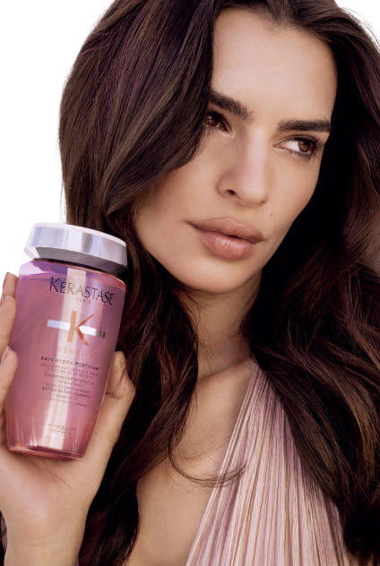 Donna che tiene una bottiglia trasparente rosa di Kerastase Genesis Bain Hydra-Fortifiant, che presenta il prodotto.