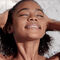 A young woman with curly hair smiles gently while washing her hair in the shower, water glistening on her skin against a marble background.