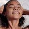 A young woman with curly hair smiles gently while washing her hair in the shower, water glistening on her skin against a marble background.