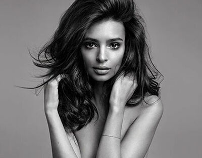 A black and white portrait of a woman with long, wavy hair, looking directly at the camera with a soft expression. She is holding her hands near her shoulders.