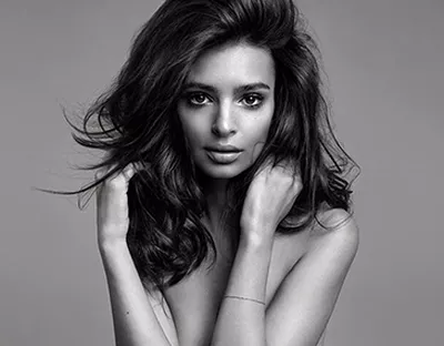 A black and white portrait of a woman with long, wavy hair, looking directly at the camera with a soft expression. She is holding her hands near her shoulders.