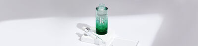 A green glass bottle of serum, partially filled with liquid, alongside a pipette, all resting on a white surface with soft shadows.