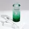 A green glass bottle of serum, partially filled with liquid, alongside a pipette, all resting on a white surface with soft shadows.