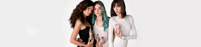 Three women pose together, holding various hair care products, with a neutral background. Each woman has a distinct hairstyle: one with curly hair, another with straight hair and teal streaks, and the third with sleek, straight hair.