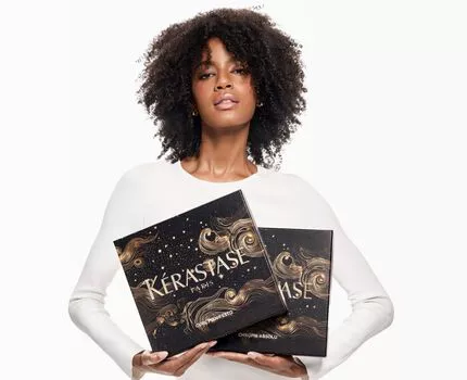 A young woman with curly hair holds two boxes with intricate designs against a white background. She is wearing a white long-sleeve shirt.