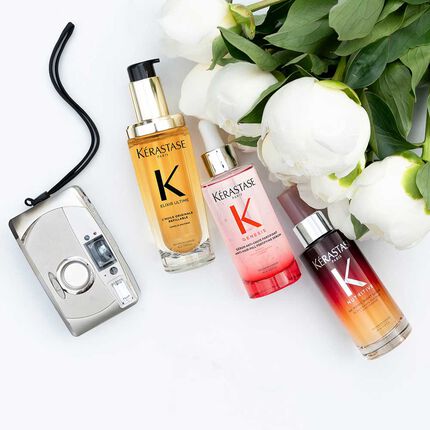 Tre flaconi di prodotti per capelli Kerastase (Elixir Ultime, Genesis e Nutritive) sono disposti su una superficie bianca accanto a una macchina fotografica vintage e a diverse peonie bianche con foglie verdi. Il flacone dorato Elixir Ultime e a sinistra con una pompa nera, il flacone rosa Genesis con un contagocce argentato e al centro, e il flacone sfumato rosso-arancio Nutritive con un contagocce argentato e a destra. La macchina fotografica argentata con il suo laccetto nero e a sinistra dei prodotti.
