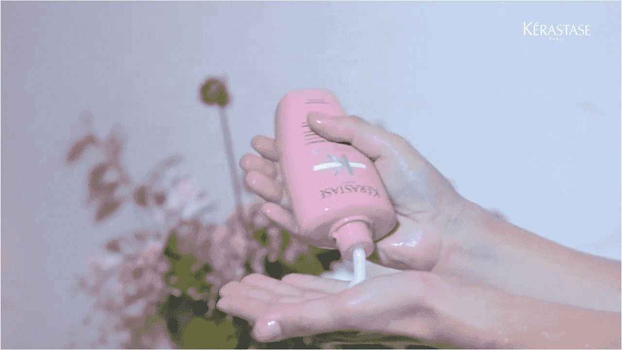 Mani che versano un prodotto per capelli Kérastase in una palma, con sfondo di fiori sfocati.