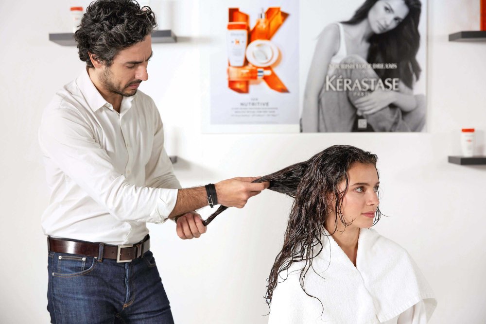 Un parrucchiere pettina i capelli di una giovane donna in un salone, circondati da prodotti Kérastase.