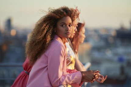 Tre donne con stili diversi, una in primo piano con capelli ricci e castani, si affacciano su una città al tramonto.